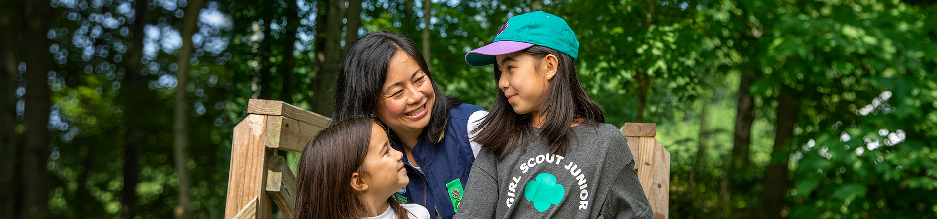  adult volunteer wearing vest with a girl scout junior (right, in baseball cap) and daisy (left) outside at a park smiling and looking at one another 